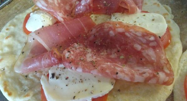 Die Fladen mit Tomaten, Mozzarella, Schinken und Salami belegen. Frisch gemahlenen Pfeffer auf den Mozzarella geben.