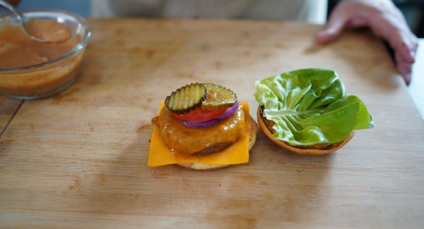 Eine Scheibe von der roten Zwiebel, Tomate und zwei Scheiben von den Gewürzgurken drauflegen. Auf der andern Seite des Brötchen kommt ein Salatblatt.